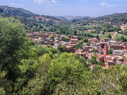 Blick über das Dorf Zaouia d’Ifrane, Lehmhäuser und ein Minarett zwischen Bäumen, dahinter die grünen Hügel des Mittleren Atlas