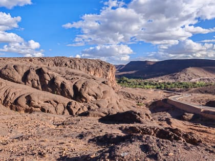 Dunkle Felsrücken vor Ouarzazate, im Tal ein grüner Streifen Palmen, dahinter die Tafelberge