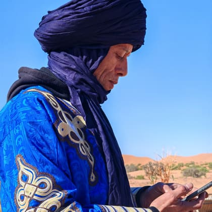 Ali im blauen Gewand mit Turban, das Telefon in der Hand, dahinter die Dünen