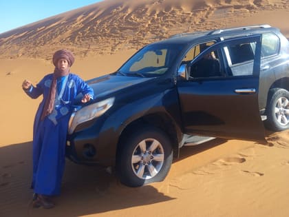 Ali im blauen Gewand neben dem Geländewagen am Fuß einer Düne